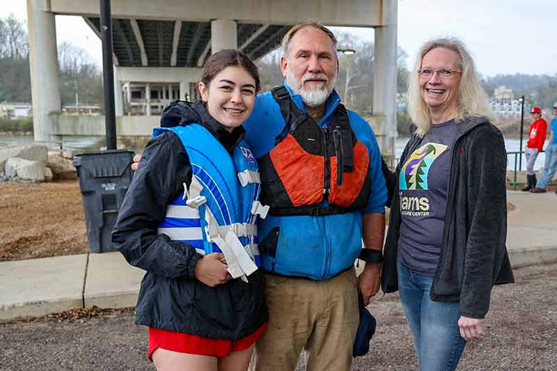 Man and woman in life jackets next to another woman, standing under a bridge