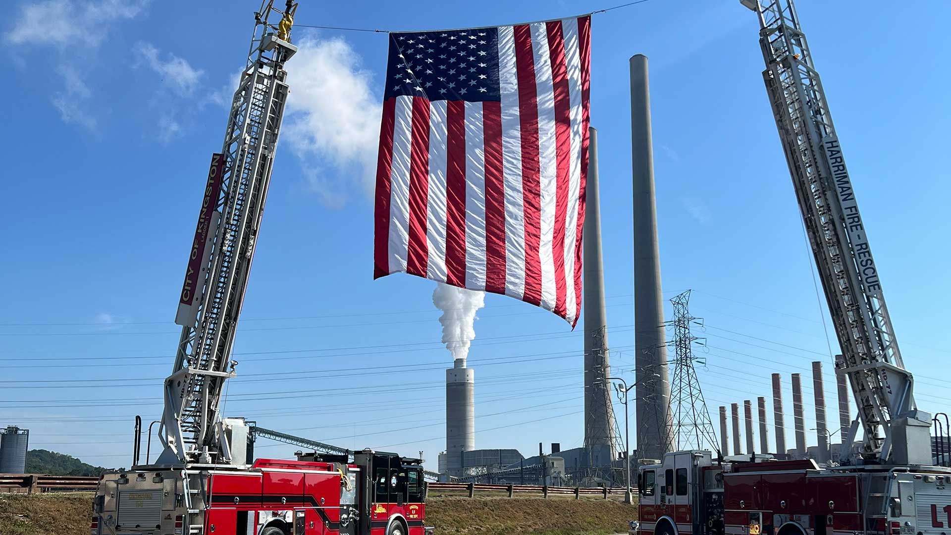 An American flag is suspended in the air by two ladder trucks with Kingston Fossil Plant in the background.