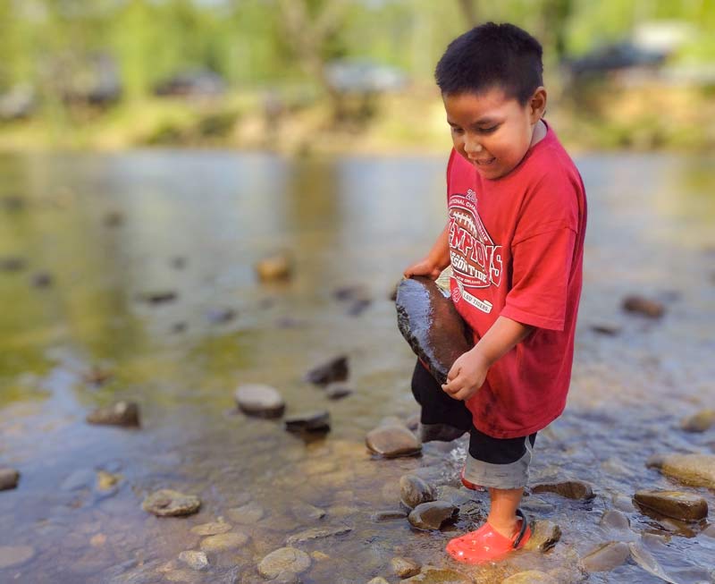 A small boy carries a large rock while walking through shallows at the Oconaluftee River