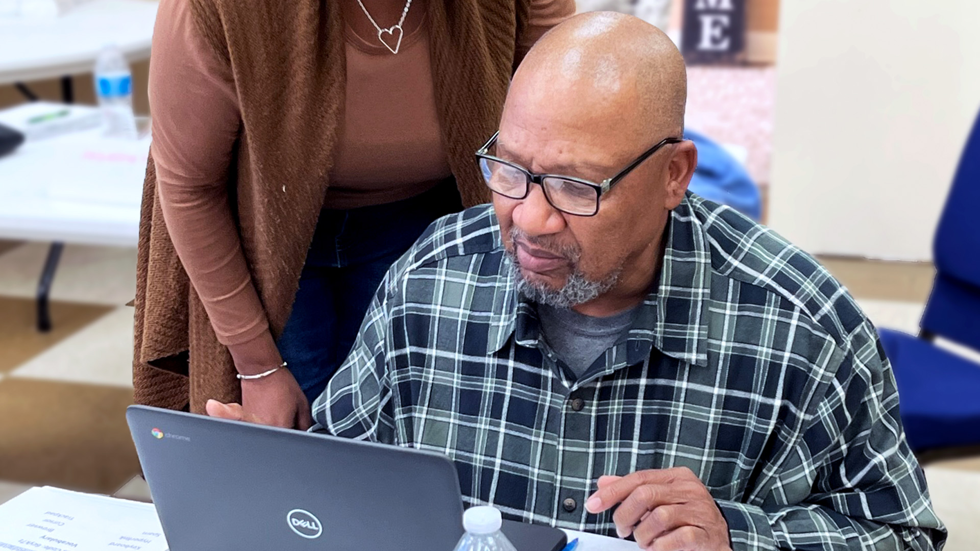 Man wearing glasses using a laptop, with a woman standing beside him offering support.