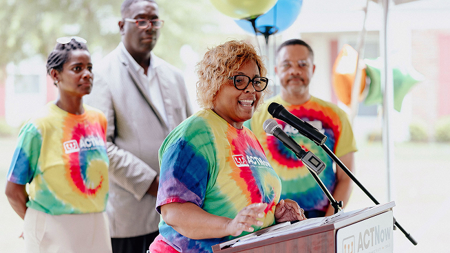 A woman speaks at a podium with others standing behind her in colorful shirts.