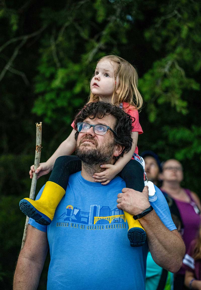 A young girl rides on a man’s shoulders as they peer up into the sky