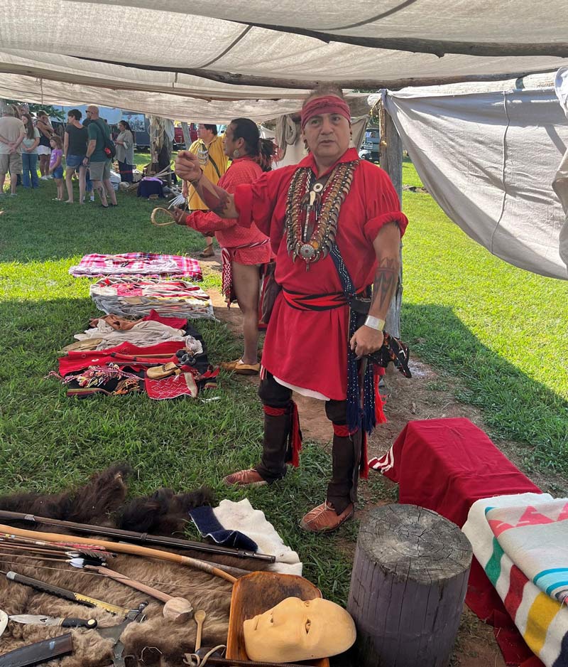 A man wearing a long, bright red shirt and brown pants stands under a white awning where a series of Native American cultural objects are on display.