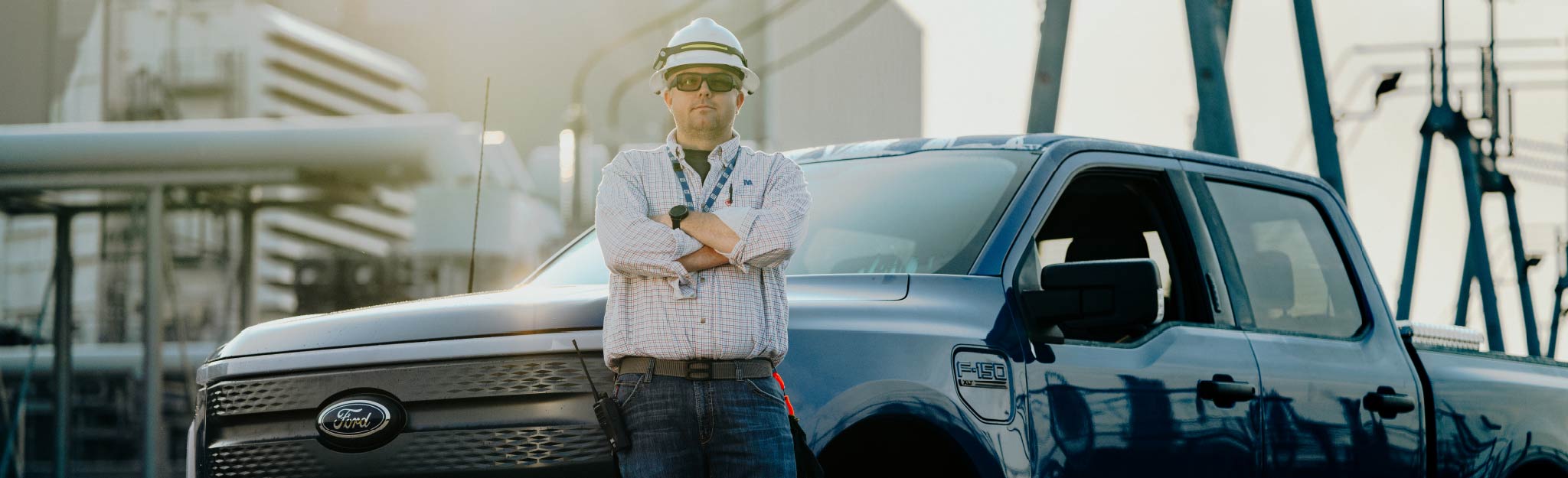 A person in a hard hat and safety glasses stands with arms crossed in front of a Ford electric truck at an industrial site