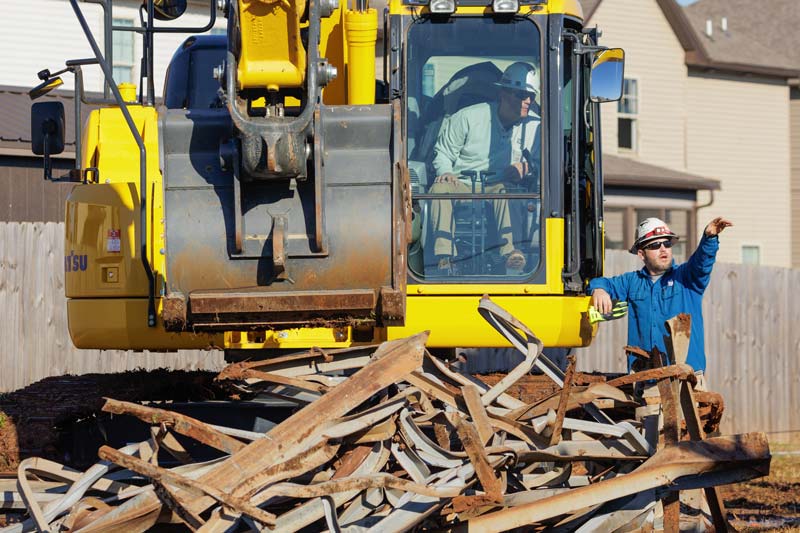 A worker signals to the operator of an excavator used to move metal debris