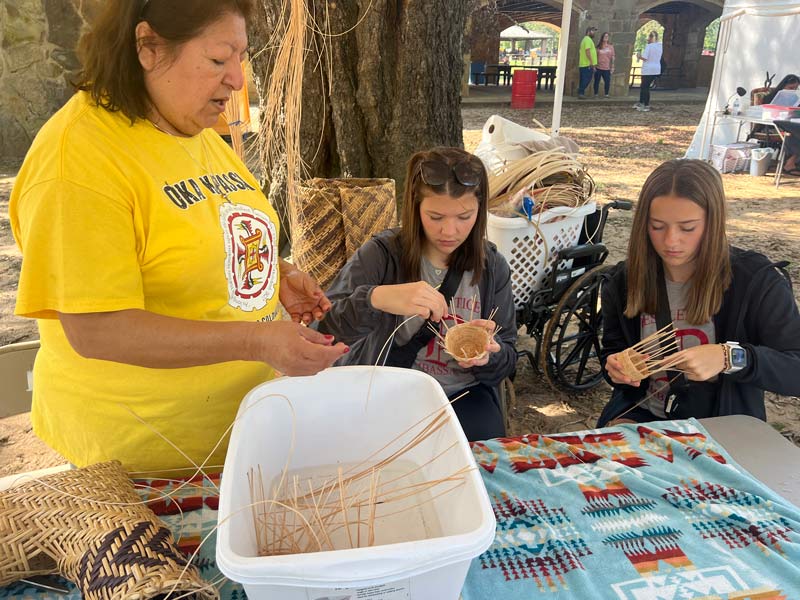 A woman in a bright yellow shirt shows two girls how to use reeds to weave a small basket