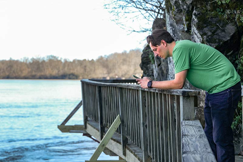 RiverLine intern leans on railing near riverfront while looking at smartphone app