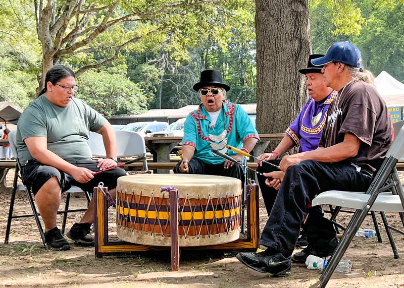 At the Oka Kapassa Festival, four men sit close to a large Native American style drum as they tap brightly colored drumsticks against it