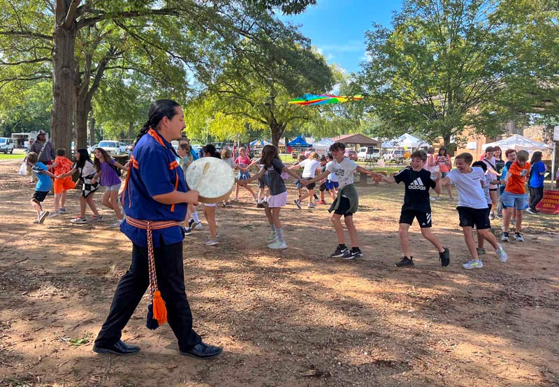 An adult strikes a handheld drum as a large group of students link hands and dance at the Oka Kapassa Native American Festival