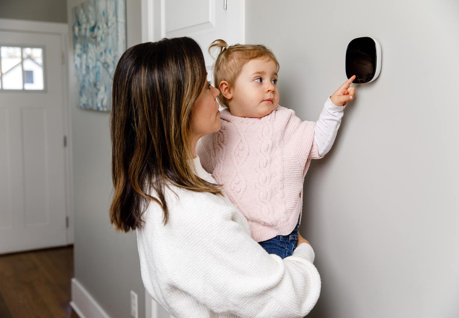 A mother holding her toddler in a pink sweater as the child reaches toward a wall thermostat