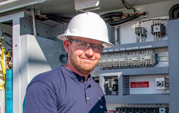 Jonathan McCullough wearing a hard hat and safety glasses, standing in front of an industrial electrical equipment