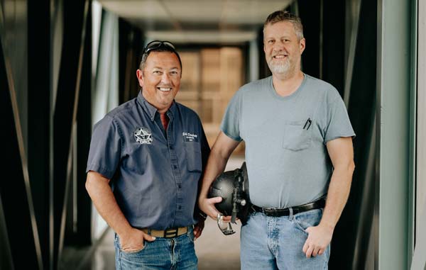 Two men stand in a corridor, smiling at the camera